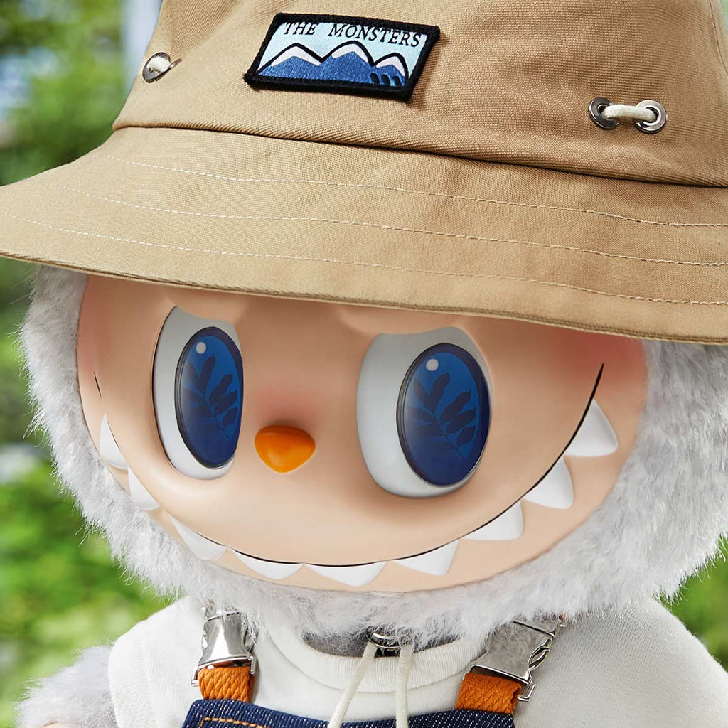 POP MART Labubu with a tan hat and blue eyes, wearing suspenders, against a blurred green background.