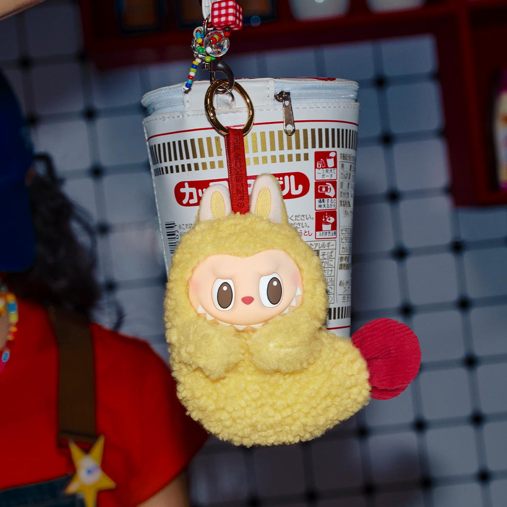 Yellow POPMART the MONSTERS Wacky Mart Series Earphone keychain with red accents hanging from a cup holder against a grid background