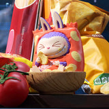 Pop Mart Labubu in a red outfit with bunny ears sitting in a wooden bowl surrounded by snacks and a tomato.
