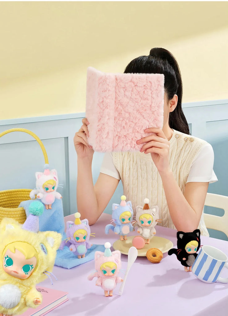 Person holding a pink POP MART Baby Molly Pocket Friends Series in front of their face with various small figurines on a table.