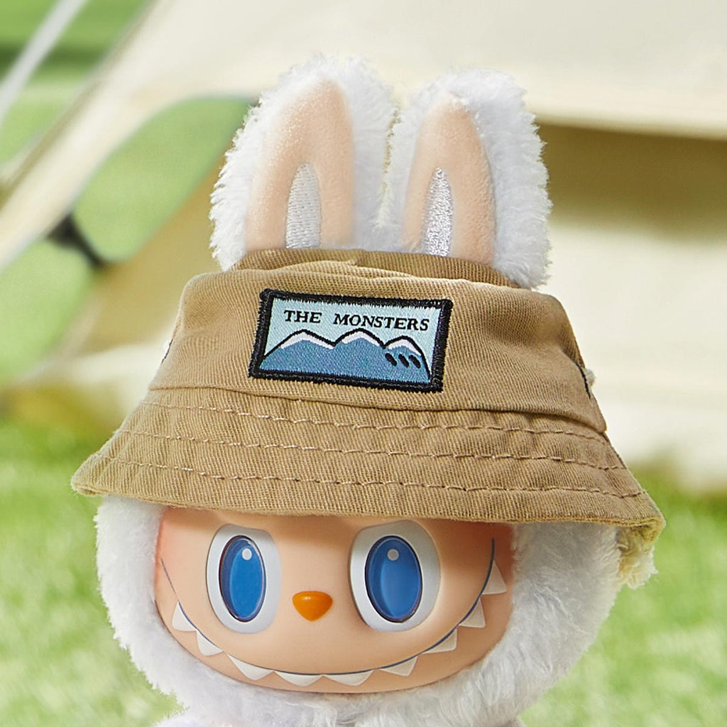 Plush Pop Mart the Labubu Monsters with a tan hat featuring a 'The Monsters' logo, set against a blurred natural background.