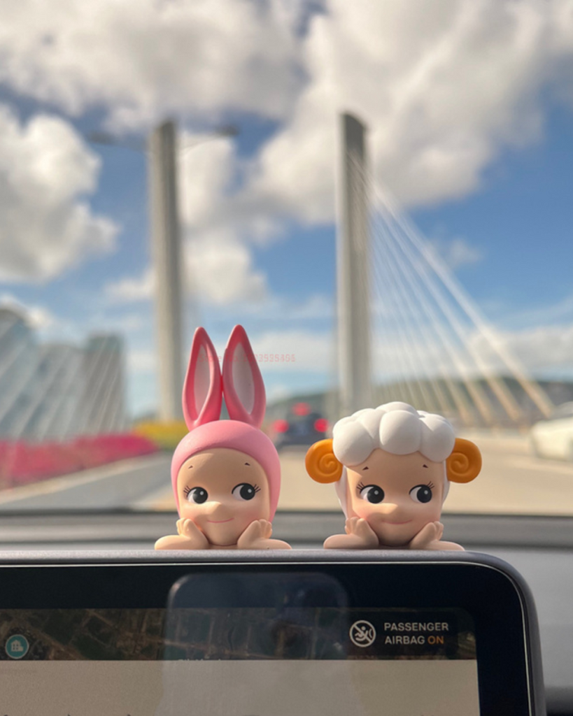 Two Sonny Angels Animal Hipper on a car dashboard with a blurred cityscape in the background.
