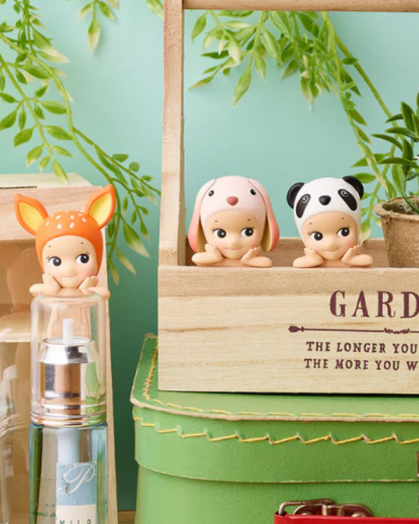 Three Sonny Angels Animal Hipper in a wooden crate with plants and a green container in the background.