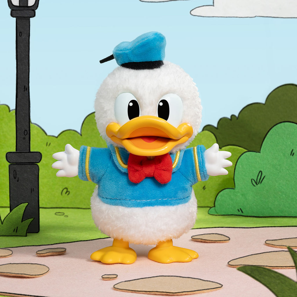 Plush toy of a cartoon character with a blue outfit and yellow shoes, standing on a path with green bushes and a lamp post in the background.