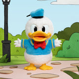 Plush toy of a cartoon character with a blue outfit and yellow shoes, standing on a path with green bushes and a lamp post in the background.